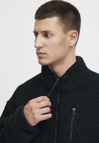 Dark green fleece jacket with a high collar, zippered front, and contrasting black zip details. Soft texture, relaxed fit, and oversized sleeves.