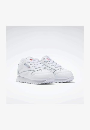 Vita atletiska skor tillverkade av syntetiskt läder med en strukturerad yta, rund tå och ett zigzag-mönster på sulan. Funktioner inkluderar snörning och logotyp.