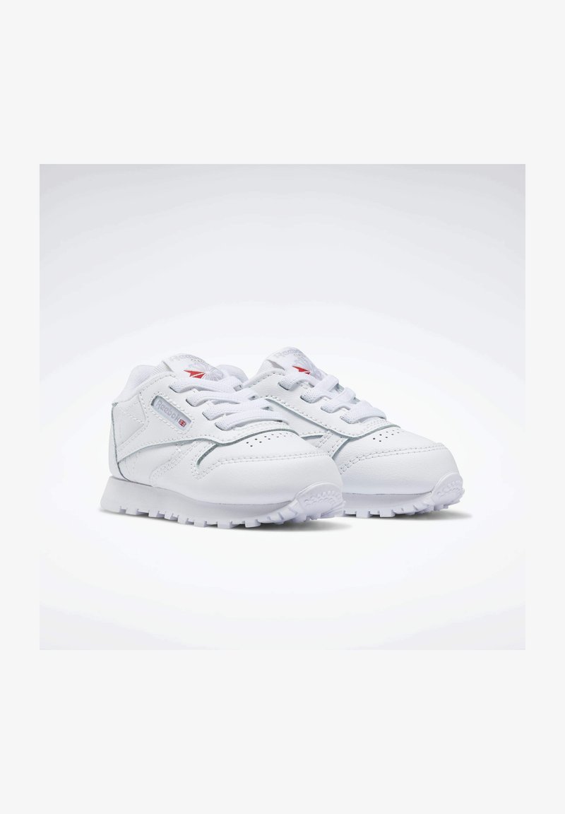 Sapatilhas brancas feitas de couro sintético com uma superfície texturizada, biqueira redonda e padrão de sola zigzag. Apresentam um design com atacadores e logótipo.