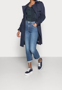Impermeabile blu marina sopra una maglietta verde a fantasia, abbinato a jeans in denim arrotolati e sneakers blu marina con lacci bianchi e suola bianca spessa.