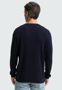Mann mit kurzen dunklen Haaren, der einen marineblauen Langarmpullover und hellblaue Jeans trägt, mit dem Rücken zu einem einfarbigen, hellgrauen Hintergrund.