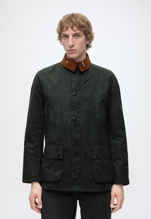 Junger Mann mit hellen, welligen Haaren trägt eine dunkelgrüne Jacke mit braunem Cordkragen, Knopfverschluss vorne und zwei Klappentaschen.