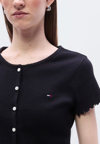 Γυναίκα που φοράει ένα μαύρο ριμπαριστό τοπ με κοντά μανίκια, λευκά κουμπιά και ένα μικρό κόκκινο, λευκό και μπλε λογότυπο στο στήθος.