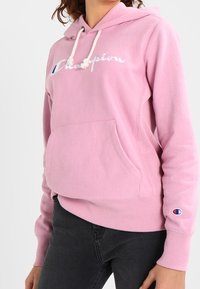 Roze hoodie met een voorzak, trekkoordsluiting en geborduurd "Champion" logo op de borst en mouw. Katoenmengsel stof.