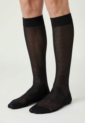 Benen met zwarte, halfdoorzichtige kniekousen met verstevigde tenen en boordjes, staand op een lichte ondergrond.