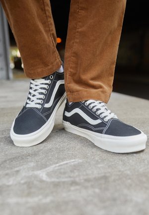 Sapatilhas de lona pretas com detalhes brancos, apresentando um design clássico, bico redondo e atacadores planos, usadas com calças de corduroy castanhas.