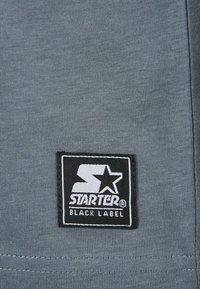 Sivá tkanina s ryhovanou textúrou má čiernu štvorcovú nálepku s bielym textom a logom hviezdy, ktorá znie "STARTER BLACK LABEL."