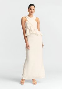 Robe longue crème avec un superposé à volants au niveau du buste et des bretelles croisées. Texture lisse, design longueur sol, associée à des sandales légères.