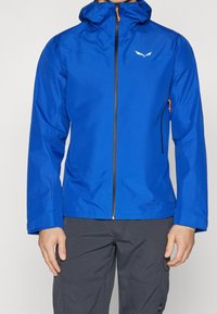 Blå vattentät jacka med huva, främre dragkedja och orange detaljer. Har en slimmad design och elastiska ärmslut, tillverkad av lätt material.