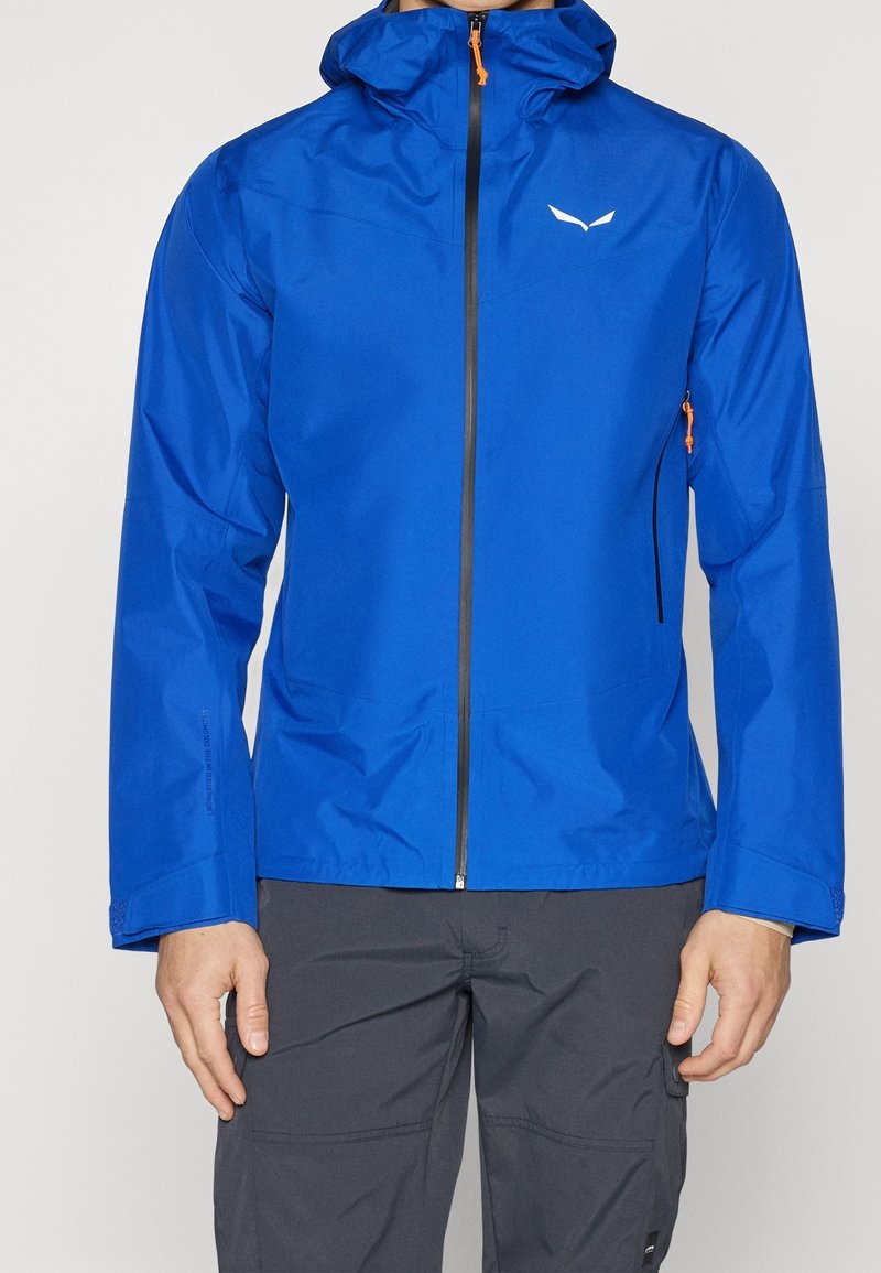 Blå vattentät jacka med huva, främre dragkedja och orange detaljer. Har en slimmad design och elastiska ärmslut, tillverkad av lätt material.