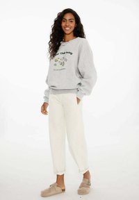 Sivá mikina s zeleným grafickým textom a ilustráciami, v kombinácii s bielymi voľnými nohavicami a béžovými slip-on topánkami. Kučeravé vlasy, v stoji.