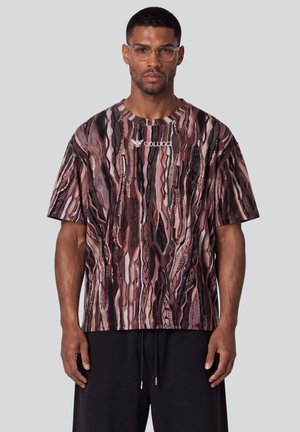 Hombre con gafas rectangulares transparentes y una camiseta holgada marrón con estampado y el logo "Colucci", combinada con pantalones negros sobre fondo liso.