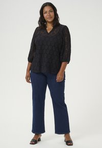 Blouse en dentelle noire à manches trois-quarts, avec un décolleté en V, portée sur un jean large bleu et des sandales à talons noires.