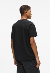 Vīrietis ar īsiem lokainiem matiem, ģērbts vienkrāsainā melnā T-kreklā un tumšos džinsos, stāv ar muguru pret balto fona.