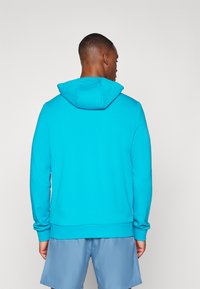 Tirkizna hoodie s prednjim kangaroo džepom, žbukanih manžeta i poruba. Ima prostranu kapuljaču i mekanu tkaninu. Nositi s plavim šortsevima.