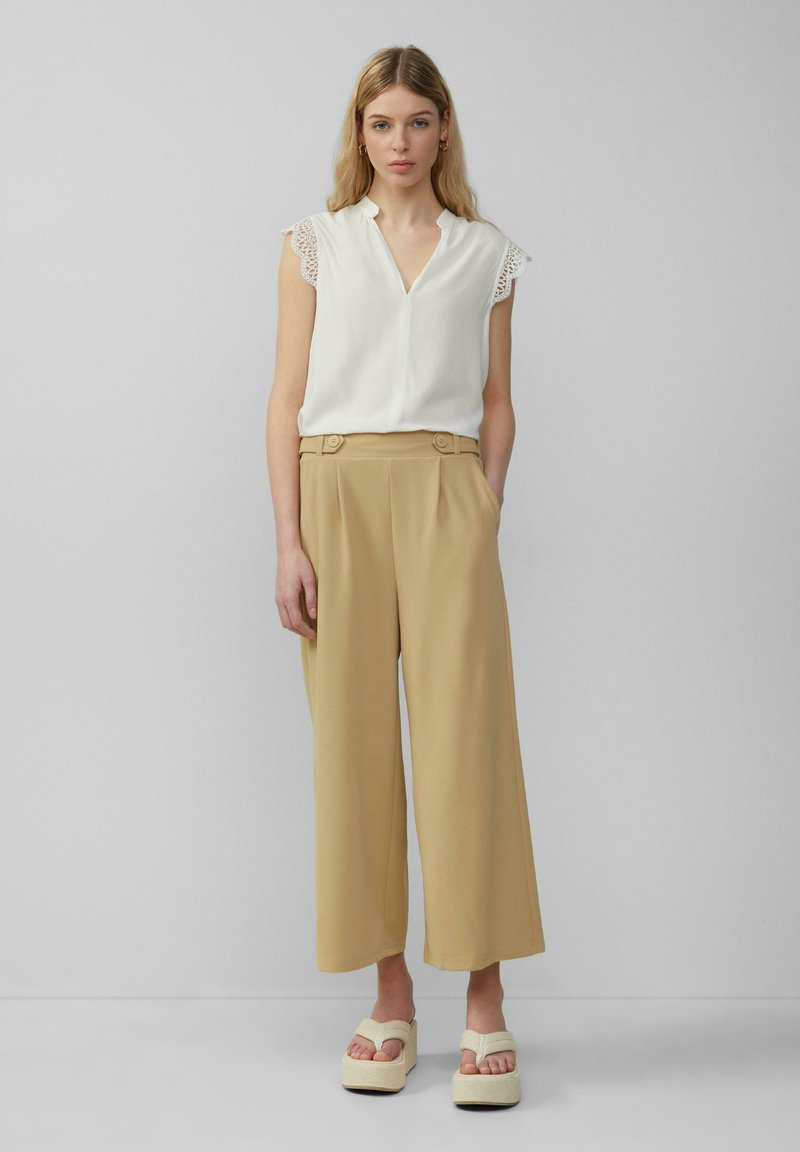 Witte blouse met kantafwerking, gecombineerd met hoog-gewaste beige wijde broek met plooien aan de voorkant en knoopdetails, gedragen met crèmekleurige platform sandalen.