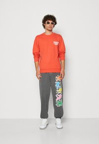 Felpa arancione con logo sul petto, abbinata a pantaloni della tuta grigi decorati con grafiche colorate. Sneakers bianche completano il look.