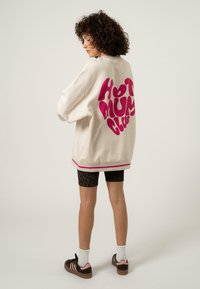 Világos bézs, oversized pulóver, rajta "HOT MUM CLUB" felirattal élénk rózsaszínben, bordás mandzsettákkal és alsó szegéllyel, rózsaszín díszítéssel. Fekete leopárdmintás sorttal viselve.