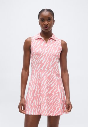 Kobieta ubrana w bezrękawnikową, różowo-białą sukienkę w kołnierzykiem i wzorem, z rozkloszowanym dołem, stojąca na tle jednolitego, jasnego tła.