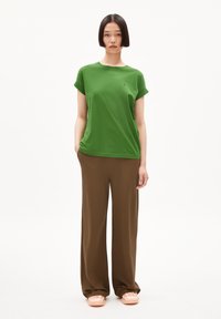 Camiseta de algodón verde con mangas enrolladas, combinada con pantalones de pierna ancha marrones. La modelo lleva calzado rosa claro y está de pie contra un fondo blanco.