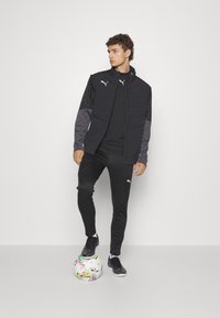 Schwarze Sportjacke mit gepolsterter Vorderseite und grauen Ärmeln, kombiniert mit einem schwarzen Top und Trainingshosen, während sie auf einem bunten Fußball steht.