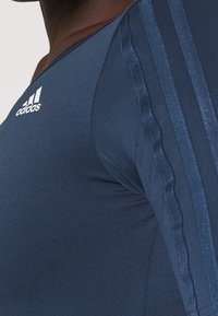 Haut de sport bleu marine avec une texture lisse, comportant trois rayures verticales le long des manches et un logo Adidas sur la poitrine.