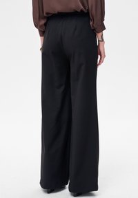 Czarne spodnie o szerokich nogawkach z elastycznym pasem, gładką teksturą i luźnym krojem, widoczne od tyłu, ukazujące eleganckie linie i fałdy.