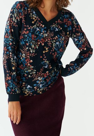 Femme portant un chemisier à manches longues à motif floral sombre avec des feuilles bleues, rouges et beiges, assorti à une jupe violette foncée.