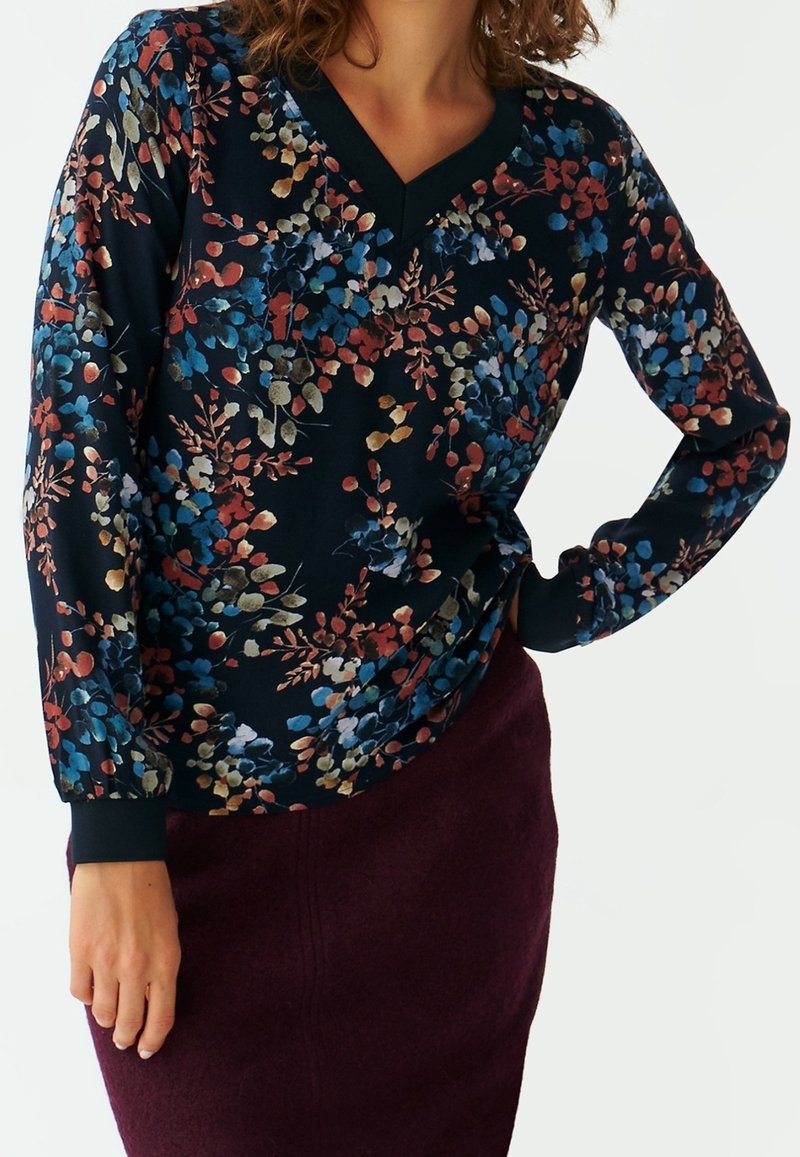 Femme portant un chemisier à manches longues à motif floral sombre avec des feuilles bleues, rouges et beiges, assorti à une jupe violette foncée.