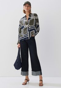 Patroonzijde shirt in groen en marineblauw met geometrische vormen, gecombineerd met wijde donkere spijkerbroeken met contrasterende manchetten en zwarte sandalen.