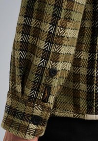 Monivärinen, herringbone-kuvioinen takki vihreänä, ruskeana ja mustana. Huomattava napitus ja kankaan rakenne näkyvissä.