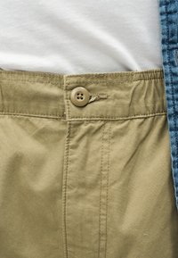 Κοντινό πλάνο στη μέση χακί παντελονιού με κουμπί κλειστό πάνω σε λευκό ύφασμα και ένδυμα από τζιν στη δεξιά άκρη.