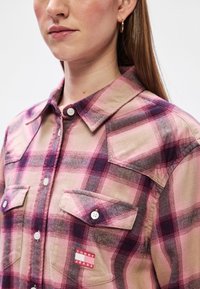 Camisa xadrez em tons de rosa e roxo, com fecho de botões na parte da frente, dois bolsos no peito e um patch da marca. Material em algodão.