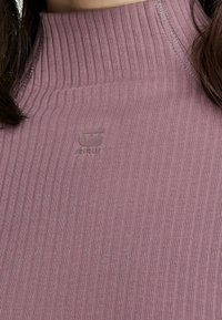 Ribbad polokrage-tröja i dämpad mauve, med en subtil logotyp och textilstruktur. Detaljer inkluderar en hög krage och rena sömmar.