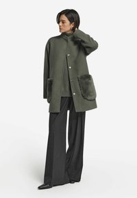 Grön ullblandad kappa med fuskpälsfickor i sidorna, knappar och en mandarinkrage, tillsammans med vida byxor och spetsiga lägenheter.