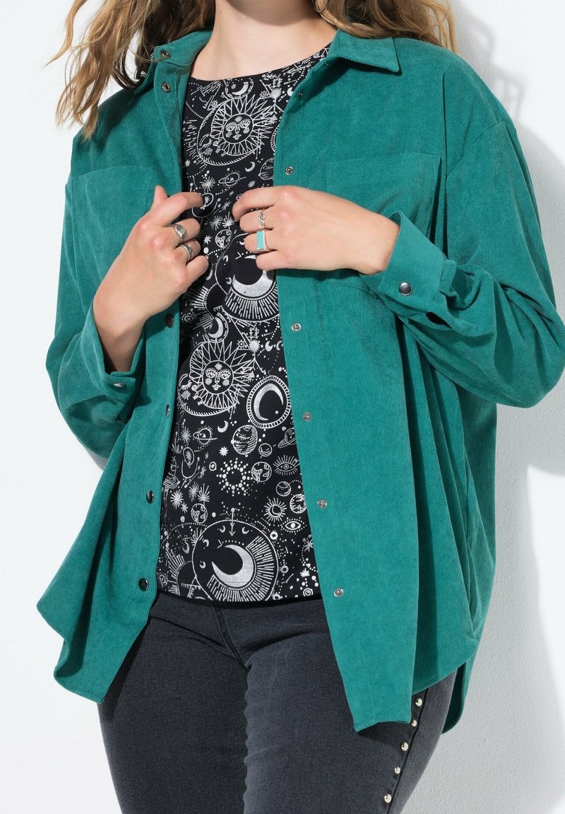 Chemise en velours côtelé teal avec des boutons, superposée sur un haut noir à motifs avec des designs célestes. Portée avec un jean noir et des clous décoratifs.