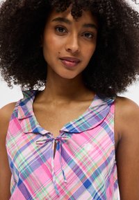 Γυναίκα με σγουρά μαλλιά, που φοράει αμάνικη μπλούζα σε ροζ, μωβ και πράσινο καρώ με μικρό φιόγκο στο ντεκολτέ, χαμογελάει απαλά.