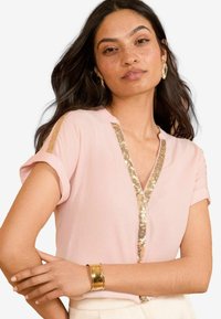 Pale pink blouse with short rolled sleeves, featuring a gold sequin trim along the neckline and front opening. Smooth fabric texture.