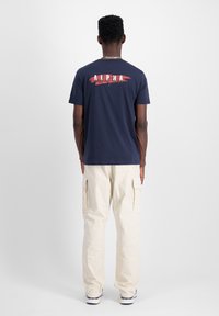 Marineblaues Baumwoll-T-Shirt mit einem roten und weißen "ALPHA"-Grafik auf der Rückseite. Kombiniert mit cremefarbenen Cargo-Hosen und schwarzen Sneakern.