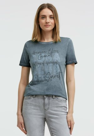 Junge Frau mit glattem braunem Haar trägt ein ausgewaschenes blaues Grafik-T-Shirt und hellgraue Jeans, steht vor einem einfachen hellen Hintergrund.