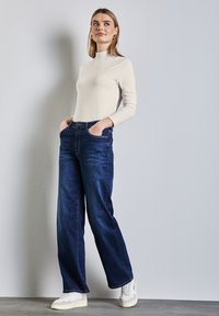 Ljus beige långärmad topp i kombination med mörkblå vida jeans och vita sneakers. Slät tygkvalitet med en figurnära design; byxorna har ett klassiskt denimutseende.