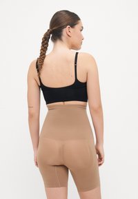 Shorts modellanti a vita alta beige con una superficie liscia, progettati per fornire supporto e una silhouette affusolata. Reggiseno nero indossato sopra.