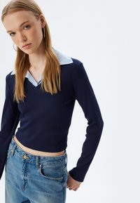 Dunkelblaues Langarmshirt mit V-Ausschnitt und hellblaurem gestreiften Kragen, kombiniert mit hochtaillierten Jeans aus Denim. Die Textur wirkt gerippt.
