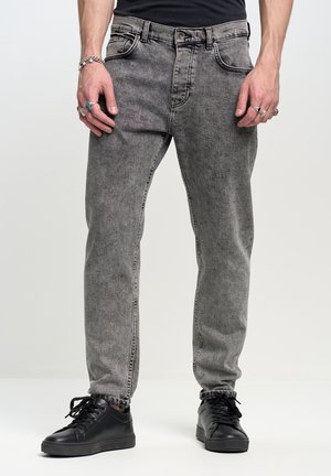 Mężczyzna w wyblakłych szarych dżinsach, czarnych trampkach, z wieloma pierścionkami i bransoletką, stojący na tle jednolitego jasnego tła.