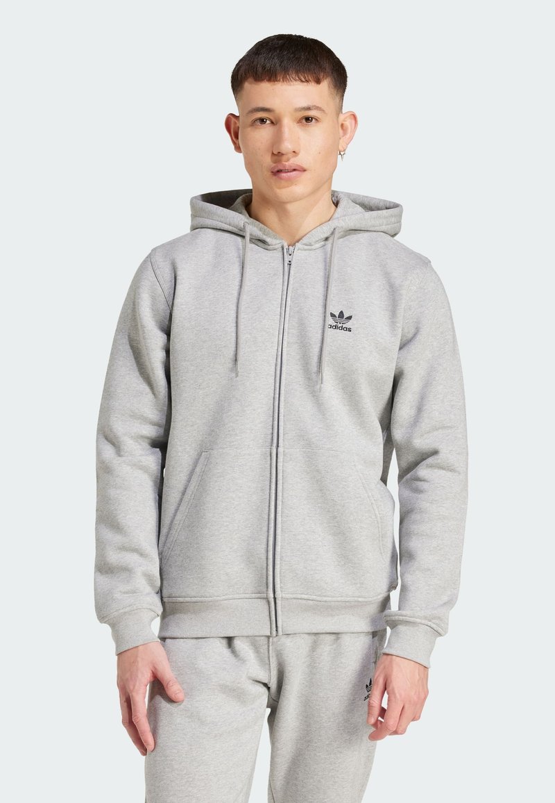 Siva zip majica s patentnim zatvaračem izrađena od meke tkanine, s prednjim patentnim zatvaračem, džepovima i malim crnim logotipom na prsima.