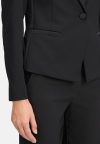 Schwarzer maßgeschneiderter Blazer mit glatter Textur, Ein-Knopf-Verschluss und seitlichen Taschen. Die Ärmel sind schmal geschnitten und das gesamte Design ist elegant.