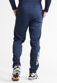 Marineblå softshell joggers med elastisk talje, tætsiddende pasform og hvidt brandlogo på siden. Båret med sort og hvidt træningssko.