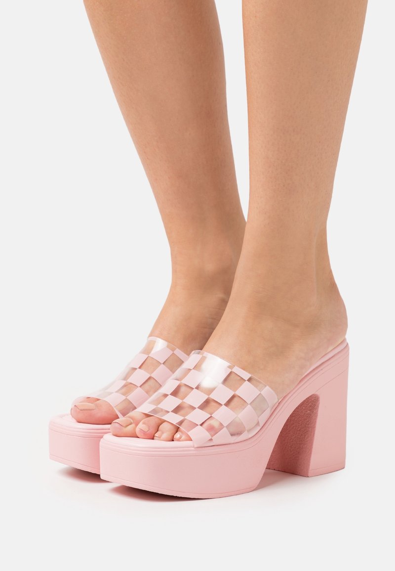 Pieds portant des talons compensés roses avec des brides transparentes à carreaux sur les orteils, présentés sur un fond clair uni.