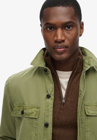 Olive zielona kurtka z guzikami, z kieszenią na piersi, noszona na brązowym swetrze zapinanym na zamek i kremowej koszuli. Gładki materiał o subtelnej fakturze.