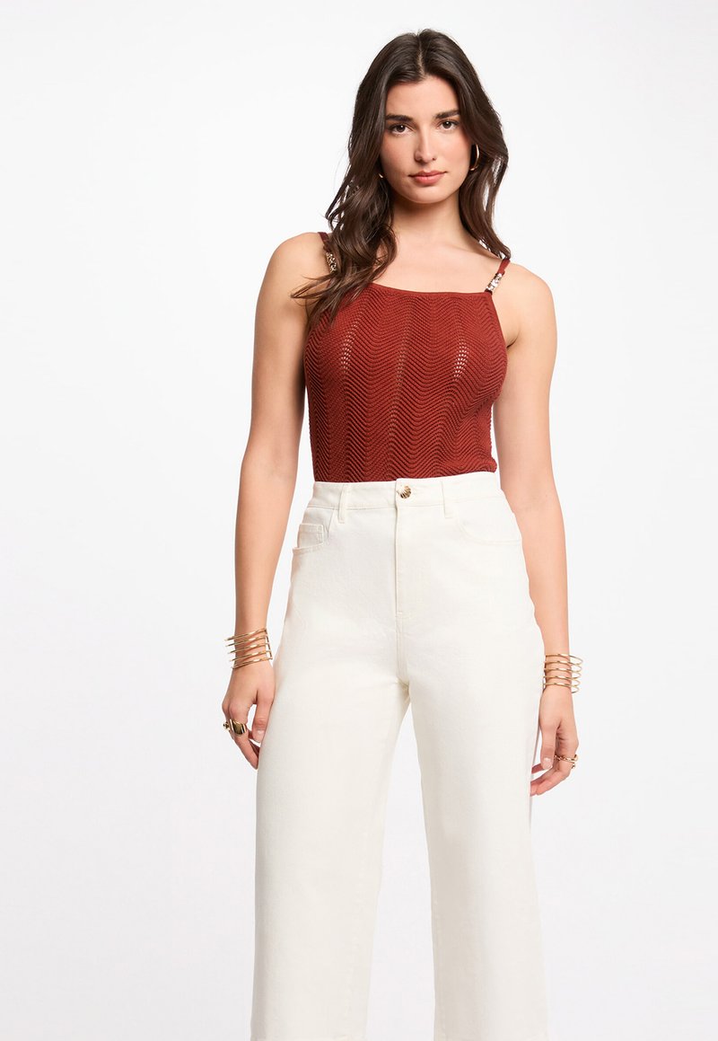 Mujer con cabello oscuro ondulado, vistiendo un top de punto sin mangas color óxido y pantalones blancos de pierna ancha de talle alto, complementada con pulseras y anillos dorados.